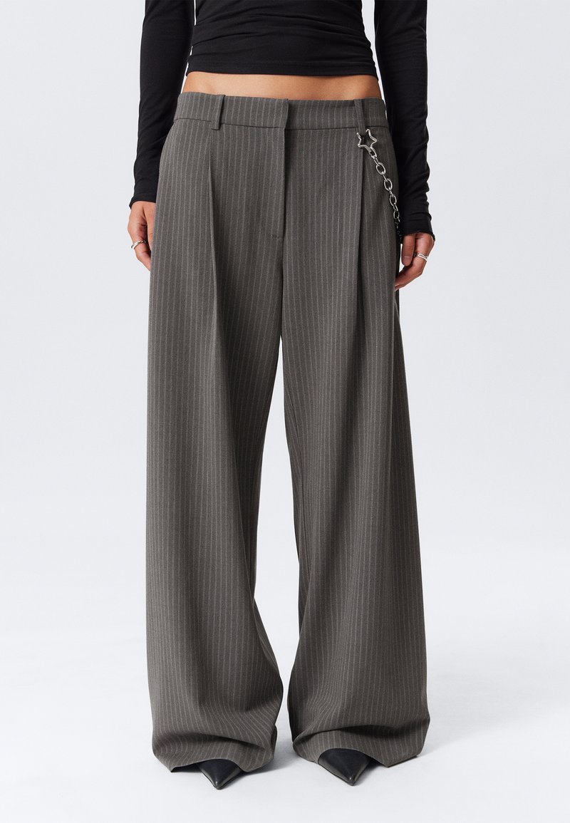 Femme portant un pantalon gris à rayures verticales, taille haute et jambes larges, avec un accessoire en chaîne argentée, associé à un haut noir à manches longues cropped.