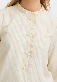 Gräddfärgad blus med vertikala blå ränder, volangkant vid halslinningen och knäppning framtill med pärlemorknappar. Mjukt tyg.