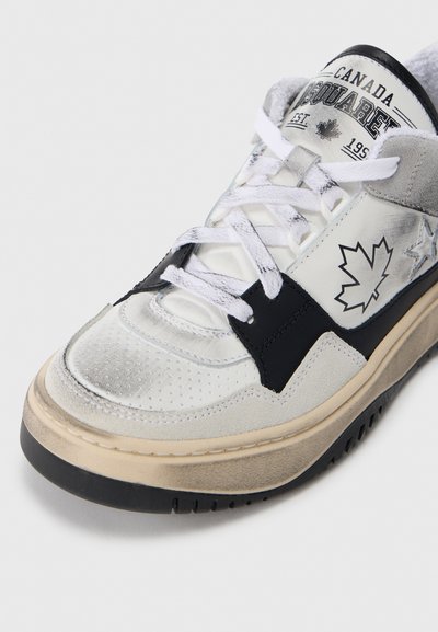 Baskets blanches et noires avec une texture perforée, une semelle robuste et un détail de feuille d'érable. Bout renforcé et lacets blancs. Semelle extérieure en caoutchouc.