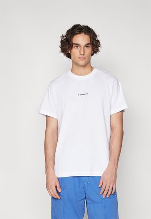 Camiseta de algodón blanca con cuello redondo y mangas cortas; presenta un gráfico de logotipo negro en el pecho. Estilizada con pantalones cortos cargo azules.