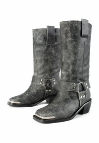 Un par de botas de vaquero de cuero negro hasta la mitad de la pantorrilla con punteras plateadas y correas decorativas con clavos metálicos alrededor de los tobillos.