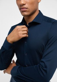 Marineblauwe longsleeve shirt van gladde stof, met een klassieke kraag en knoopsluiting met donkere knopen, getoond op de bovenlichaam van een model.