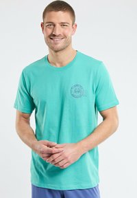 T-shirt en coton turquoise avec un col rond, des manches courtes et un logo graphique circulaire sur la poitrine gauche.