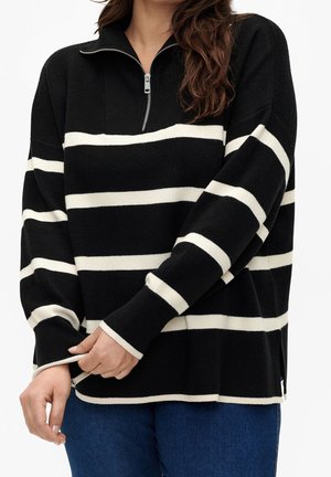 Frau trägt schwarzen Pullover mit weißen horizontalen Streifen und Frontreißverschluss, kombiniert mit blauer Jeans.