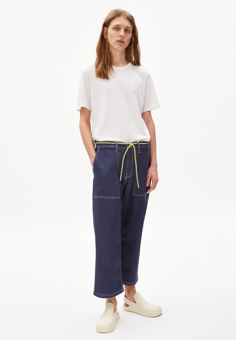 Weißes Baumwoll-T-Shirt, marineblaue Jeans-Hose mit weitem Bein und gelbem Kordelzug sowie off-white Clogs. Die Hose hat frontale Taschen und einen Kontraststich.