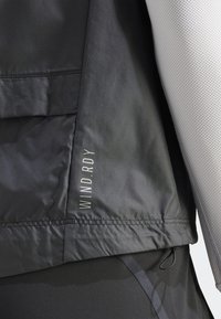 Czarna wiatrówka o gładkiej fakturze z odblaskową etykietą "WIND.RDY". Część warstwowego, sportowego stroju z czarnymi akcentami.