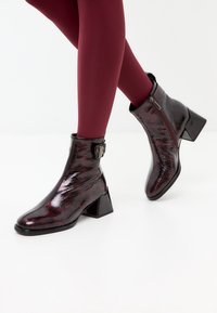 Black and burgundy patent leather ankle boots with a square toe, decorative buckle, and block heel, featuring a shiny, textured finish.