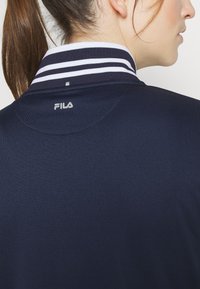Navy polo shirt featuring a striped collar in white and navy, with a small embroidered logo at the back of the neck. Smooth fabric texture.