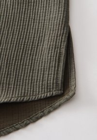 Gros plan sur un tissu texturé vert olive avec un bord plié et une petite fente latérale sur un fond clair.