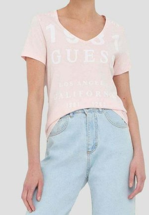 Helles rosa Baumwoll-T-Shirt mit V-Ausschnitt, bedruckt mit weißem Text "1981", "GUES" und "LOS ANGELES CALIFORNIA" in unterschiedlichen Größen. Kombiniert mit hellblauen Jeans.