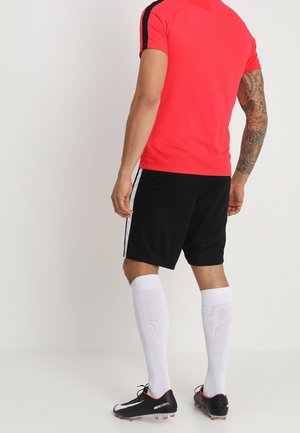 Man standing with back facing camera, wearing red short-sleeve sports shirt, black shorts with white stripes, white knee-high socks, and black cleats.