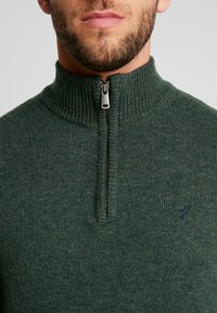 Suéter de punto verde oscuro con cuello de media cremallera, textura acanalada y un pequeño logo bordado en el pecho.