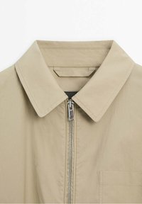 Beige jacket with a pointed collar, front zipper, and single chest pocket. Smooth fabric with minimal texture. Simple and functional design.
