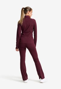 Burgundy träningsdräktset bestående av en åtsittande långärmad topp och utsvängda byxor, tillverkad av ett mjukt, elastiskt tyg med en stilren design.