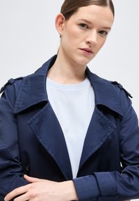 Νεαρή γυναίκα με ανοιχτόχρωμο δέρμα και καστανά μαλλιά φοράει μπλε σκούρο παλτό ύφασμα πάνω από λευκό πουκάμισο, κοιτάζοντας ελαφρώς προς την κάμερα με τα χέρια σταυρωμένα.