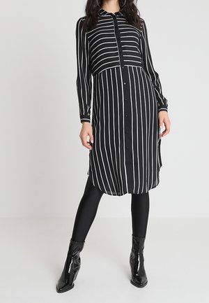 Kvinde iført en sort kjole med lodrette og vandrette hvide striber, sorte leggings og skinnende sorte ankelstøvler, stående mod en ensfarvet baggrund.