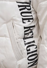 Biała kurtka puchowa z wyraźnym czarnym logo "TRUE RELIGION", charakteryzująca się teksturowanym materiałem oraz boczną kieszenią.