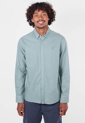 Jeune homme aux cheveux bouclés souriant, portant une chemise bleu clair boutonnée et un pantalon bleu foncé sur un fond blanc uni.