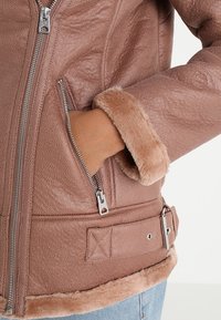 Giacca in ecopelle rosa con una finitura testurizzata, tasca con zip, colletto e orlo foderati in pelliccia, e dettagli in metallo argentato.