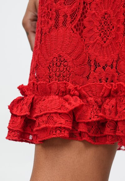 Piros csipke anyag virágmintákkal és fodros szegéllyel. A dizájn bonyolult hímzést és texturált, nyitott szemű befejezést tartalmaz.