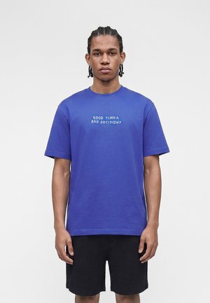 Νέος άνδρας με πλεξούδες, που φοράει μπλε μπλουζάκι με την επιγραφή «Good Times, Bad Decisions» και μαύρα σορτς, σε απλό ανοιχτό φόντο.