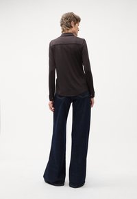 Brun langærmet skjorte med krave, parret med vide mørkeblå denimjeans. Glat tekstur, bagfra fremhæver skjortens sømme.