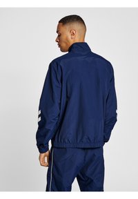 Navy blue windbreaker with a high collar, elasticized cuffs, and drawstring hem. Features white stripes on the sleeves and a smooth texture.