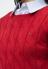 Punainen neulepusero, jossa on palmikkokuvio, ribitettävä pääntie. Kerroksittain sinivalkoraitaisen kauluspuseron päällä. Pieni logo näkyvissä.