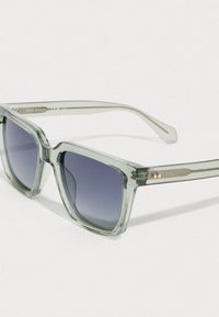 Διαφανείς ανοιχτοί πράσινοι ορθογώνιοι γυαλιά ηλίου με σκούρους κεκλιμένους φακούς που ξεκουράζονται σε μια λευκή επιφάνεια.