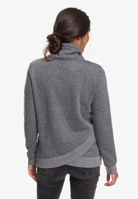 Felpa grigia con colletto alto, tessuto strutturato, orlo asimmetrico e polsini a coste. Indossata sopra jeans scuri.