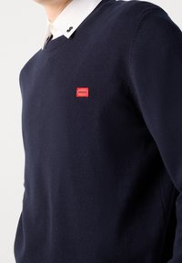 Granatowy sweter robiony na drutach z dekoltem w serek i małą czerwoną metką "HUGO" na piersi, zestawiony z białą koszulą z kołnierzykiem pod spodem.
