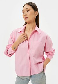 Camicia button-up rosa chiaro con tasca sul petto, vestibilità rilassata, maniche lunghe arrotolate e texture in cotone morbido. Design casual.