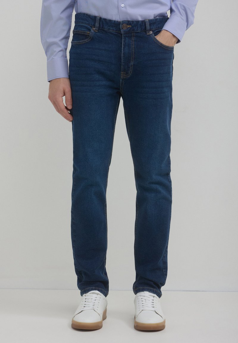 Jeans in denim blu scuro con taglio dritto, design a cinque tasche e dettagli di cucitura visibili. Abbinati a sneakers bianche.