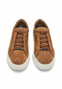 Ruskea mokkanahkakenkä, jossa on valkoiset kumipohjat, pyöreä kärki ja sävyyn sointuvat nauhat. Varustettu kohokuvioidulla brändäyksellä kielekkeessä ja sivupaneeleissa.