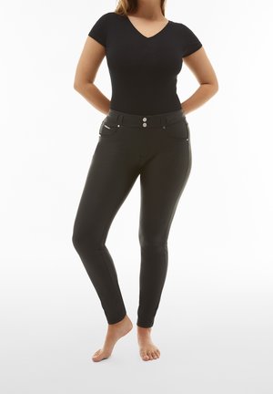 Kobieta stojąca boso, ubrana w czarny dopasowany top z krótkim rękawem i dekoltem w serek oraz czarne dopasowane spodnie z przodu z kieszeniami i podwójnym zapięciem na guziki.