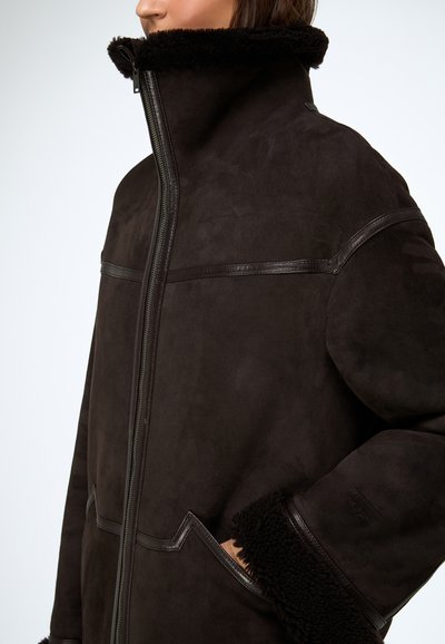Veste en daim marron avec un col montant, des accents en cuir et une doublure pelucheuse. Comprend une fermeture éclair centrale et deux poches avant.