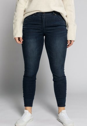 Person wearing dark blue skinny jeans, white sneakers, and a cream knit sweater standing against a plain background.