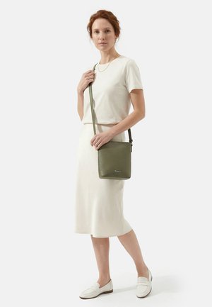 Woman wearing a white short-sleeve top and skirt, carrying an olive green crossbody bag with white loafers, standing against a plain background.