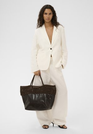 Naine, kes kannab kreemjat pintsakut ja laia säärega pükse, hoides suurt musta ja pruuni punutud käekotti, seisab ühevärvilise tausta ees.