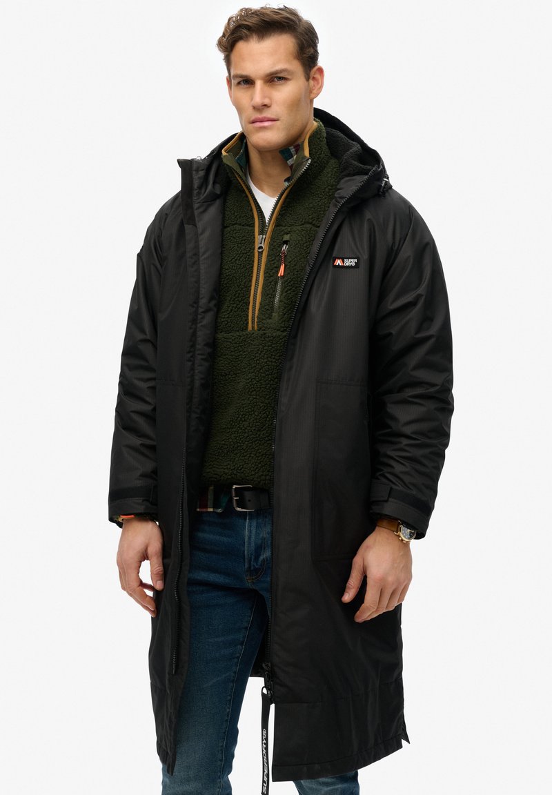 Zwarte lange parka met capuchon en ritssluiting, gelaagd over een groene fleece pullover. Voorzien van een logopatch en voorzakken.