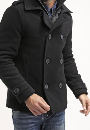 Hombre con un abrigo cruzado negro tipo pea coat sobre una camisa de mezclilla azul y vaqueros, de pie frente a un fondo liso.