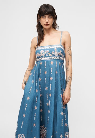 Γυναίκα με σκούρα μαλλιά και τατουάζ που φοράει ένα μπλε αμάνικο φόρεμα με σχέδια και λεπτές τιράντες, στέκεται μπροστά σε απλό φόντο.