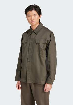 Giacca camicia verde oliva con due tasche sul petto, chiusura a bottoni e dettagli in pannelli laterali a coste. Il materiale sembra testurizzato e resistente.