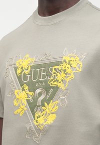Pelēka īsroku T-krekls ar zaļu trīsstūrveida logotipu, dzeltenu ziedu izšuvumu un tekstu "GUESS U.S.A. JEANS" tajā iekšpusē.