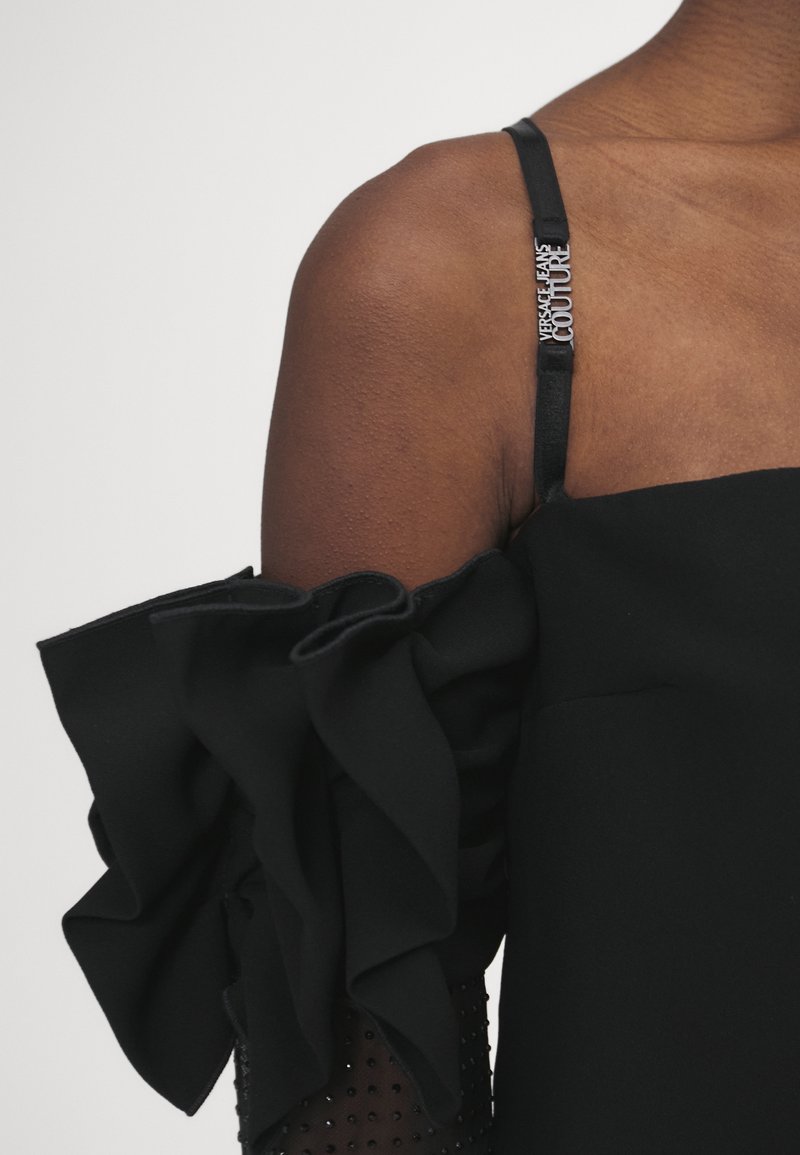 Czarna sukienka z falbaną i teksturowanym ramieniem, regulowanym paskiem z logo 'VERSACE COUTURE' oraz dopasowaną górą.