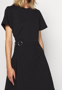 Czarna sukienka z krótkimi rękawami i okrągłym dekoltem; ma dopasowaną talię z srebrnym pierścieniem jako akcentem oraz plisami na spódnicy.
