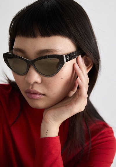 Cat-eye sunglasses in black with sparkles, gray lenses, and angular design. Worn with a red long-sleeve top. Smooth texture and distinct shape.