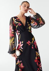 Abito nero floreale con maniche lunghe trasparenti, scollatura a V profonda e vita arricciata. Presenta vivaci motivi floreali in rosa, giallo e rosso. Tiene una borsa bianca.