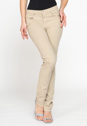 Donna che indossa pantaloni beige aderenti, top nero e tacchi aperti azzurri, in piedi con le gambe incrociate.
