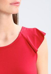 Rotes Kurzarmoberteil mit glatter Textur, rundem Ausschnitt und gefalteten Akzenten auf den Schultern.